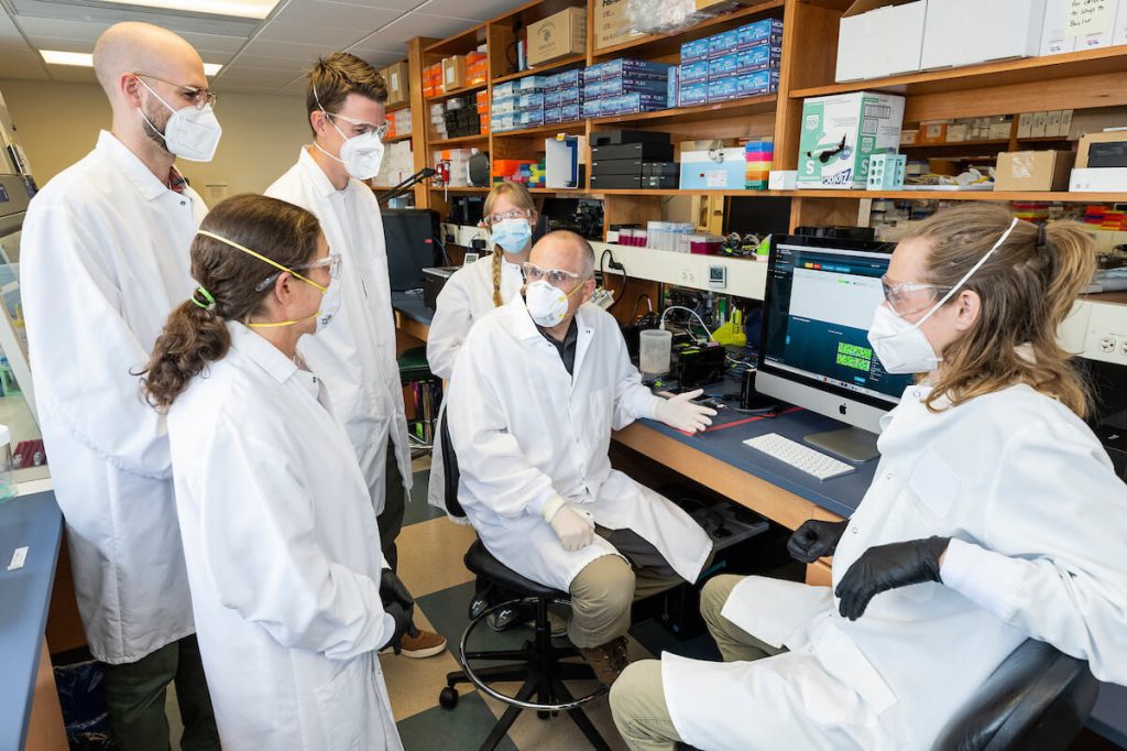 Six researchers wearing lab coats and protective masks gather around a computer workstation in a laboratory, discussing data displayed on a monitor.