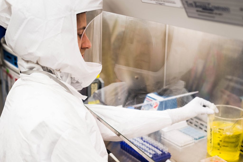 A researcher wearing a full white biohazard suit and gloves works behind a glass safety shield, handling laboratory equipment and a beaker of yellow liquid.