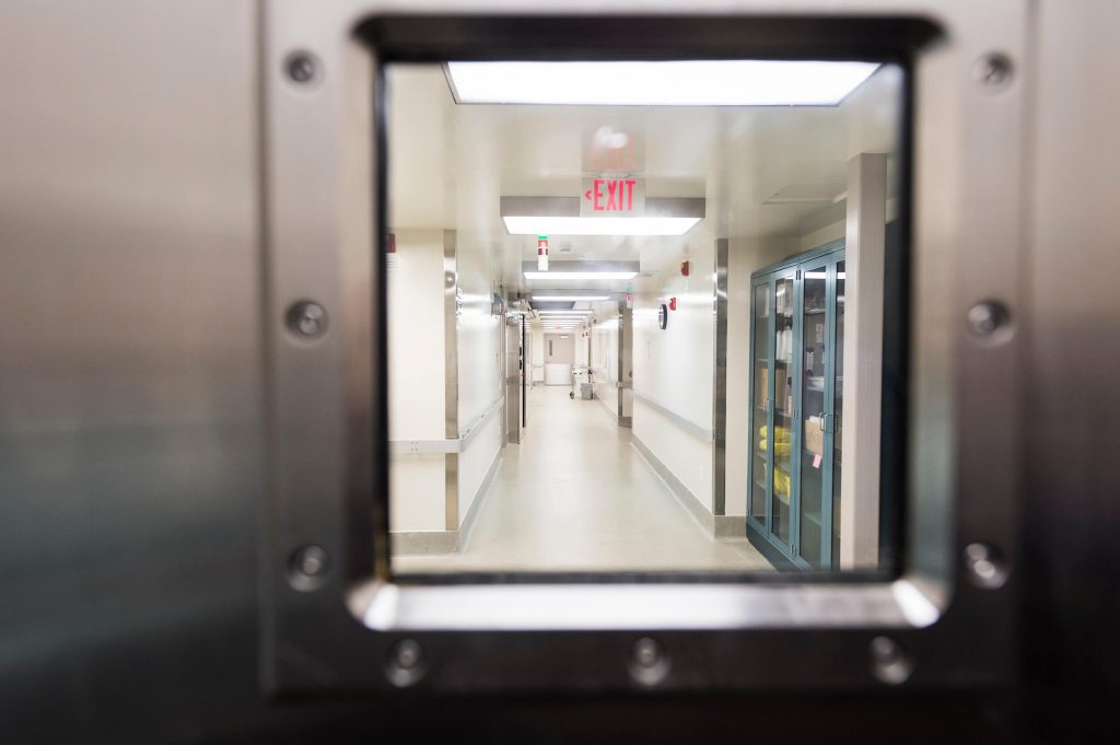 View through a small rectangular window in a metal door shows an empty, well-lit hospital corridor with an illuminated exit sign and storage cabinets along the wall.