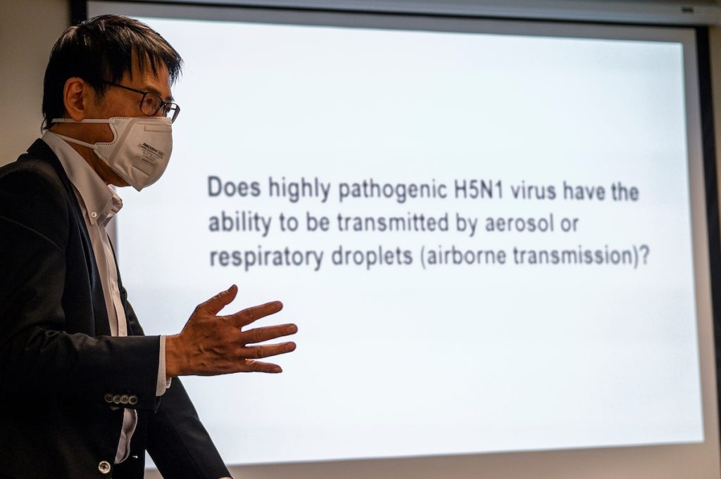 A man wearing a mask gestures as he presents in front of projected screen which reads 'Does highly pathogenic H5N1 virus have the ability to be transmitted by aerosol or respiratory droplets (airborne transmission)?