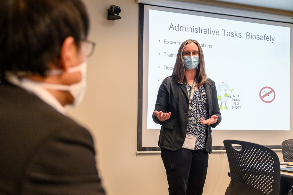 A woman wearing a face mask and glasses presents a 'Biosafety' slideshow to an audience, featuring a slide with the text 'Administrative Tasks' and a 'Get your flu shot' illustration.