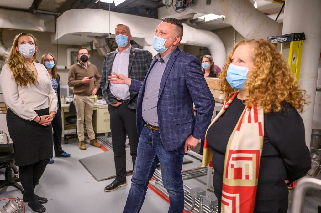 A small group of masked adults in business attire stands in a mechanical room with visible pipes and ducts, listening as one person gestures while speaking.