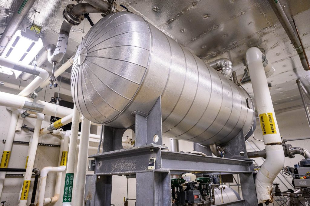 Large insulated cylindrical industrial tank mounted on a steel frame, surrounded by white pipes and valves in a mechanical room.