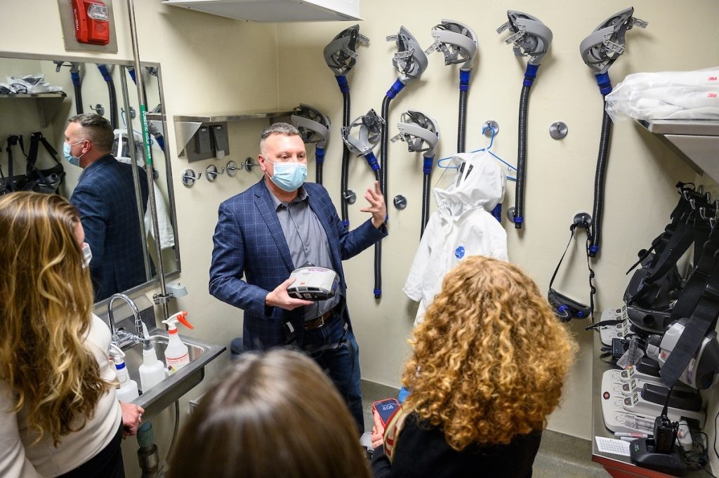 A man in a blue blazer and face mask gestures toward a piece of equipment while standing in a laboratory room surrounded by hanging respirators and protective suits.
