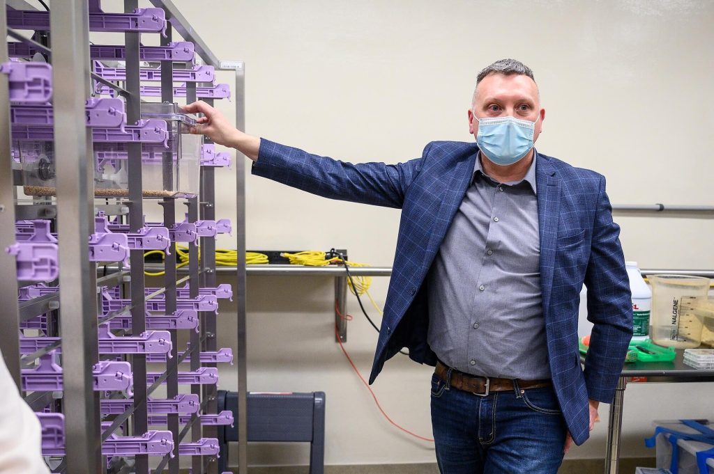 A man wearing a mask stands in a laboratory next to a tall metal rack filled with purple containers.